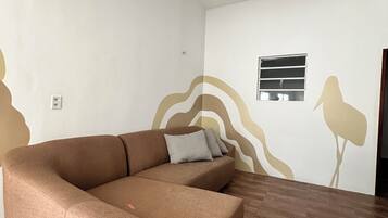 Una sala moderna con un sofá seccional marrón, una ventana pequeña con persianas y una pared decorativa con diseños abstractos.