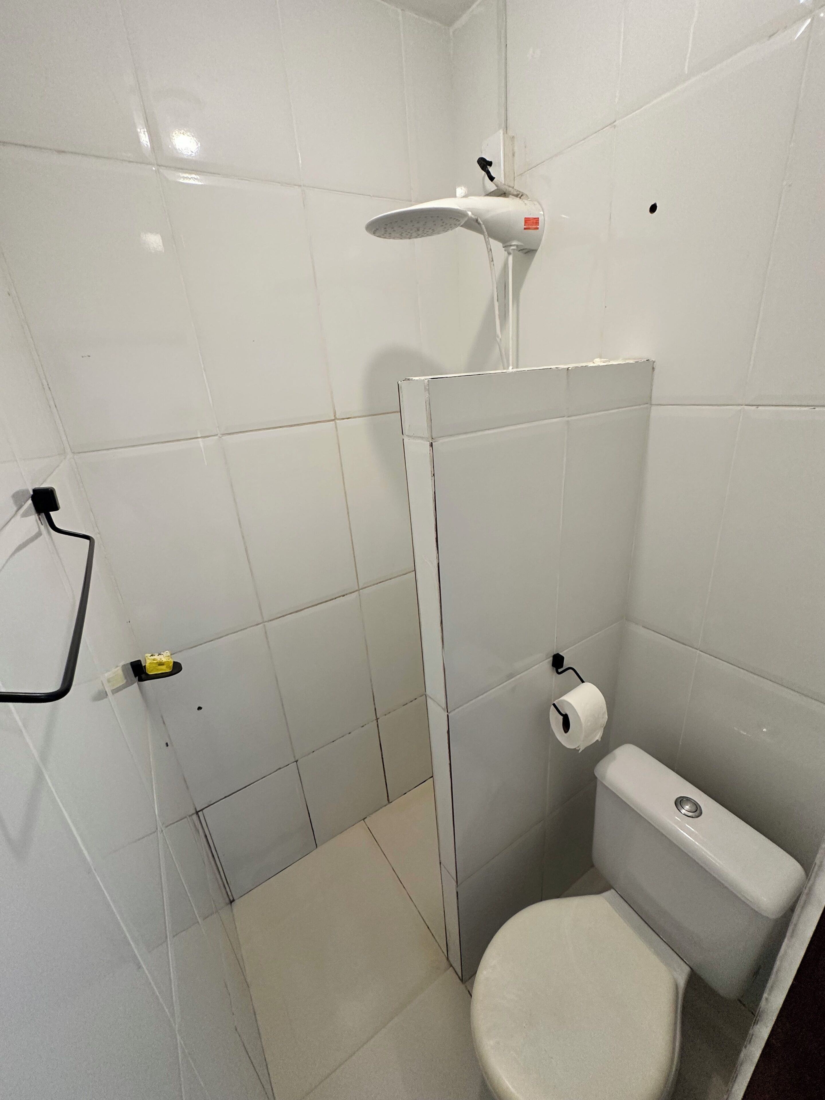 Une petite salle de bain aux murs recouverts de carreaux blancs, équipée d’une douche, de toilettes et d’un porte-serviettes.