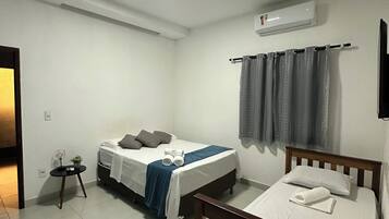 A bedroom with a bed, a bedside table, a small table with a plant, a window with curtains, and an air conditioning unit.