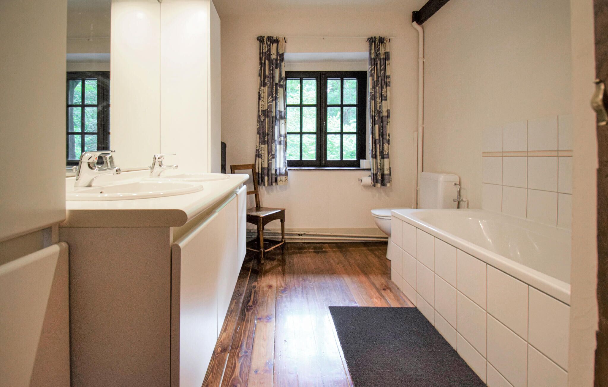 A bathroom with a wooden floor, a bathtub, a toilet, a sink, and a window with curtains.