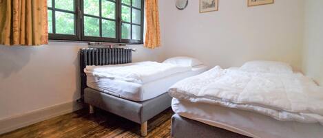 A room with two single beds, a radiator, a window with curtains, and a wall clock.