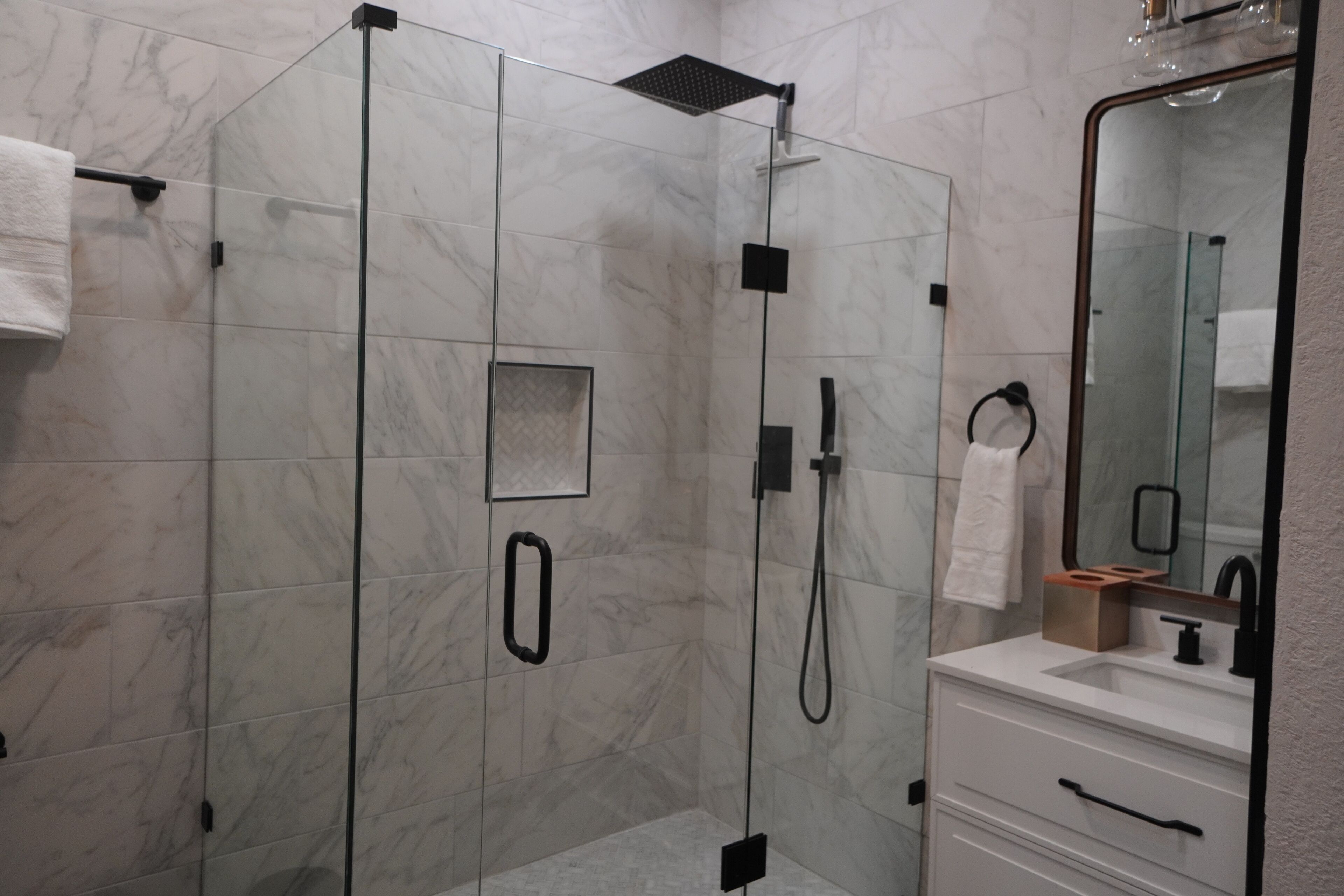A modern bathroom with a glass-enclosed shower, a white marble wall, a large mirror, and a white vanity with a sink.
