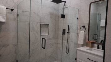 A modern bathroom with a glass-enclosed shower, a white marble wall, a large mirror, and a white vanity with a sink.