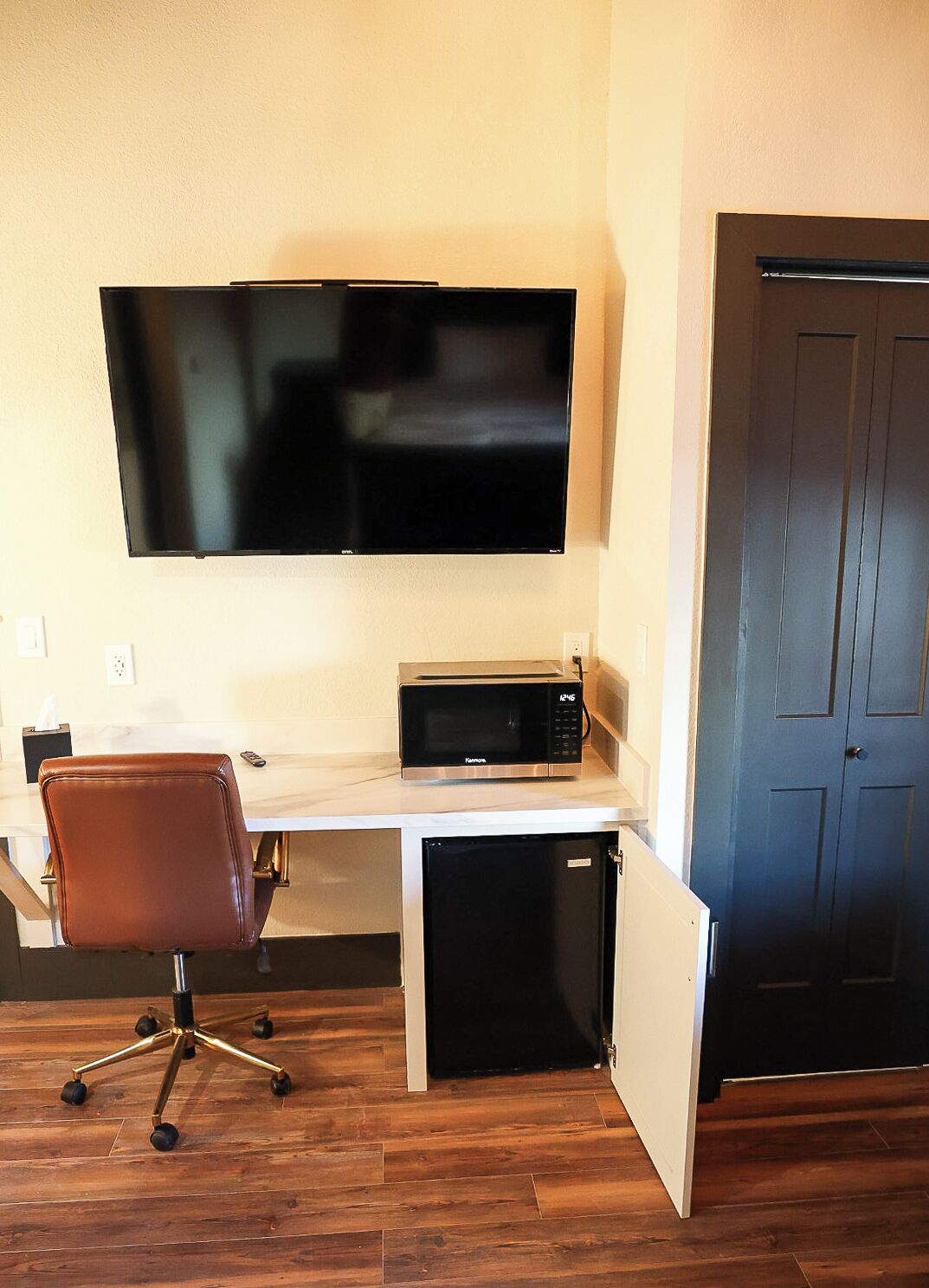 Una habitación con una televisión de pantalla plana montada en la pared, un microondas sobre una encimera, un sillón de cuero marrón y una puerta cerrada.
