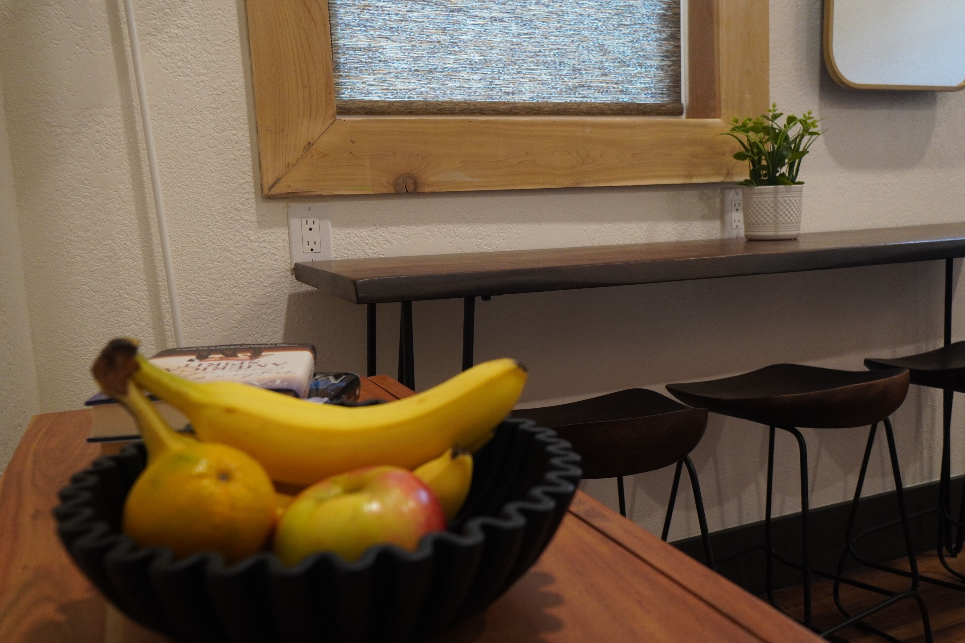 A wooden table with a black bowl containing fruits, a potted plant, and a window with a view.