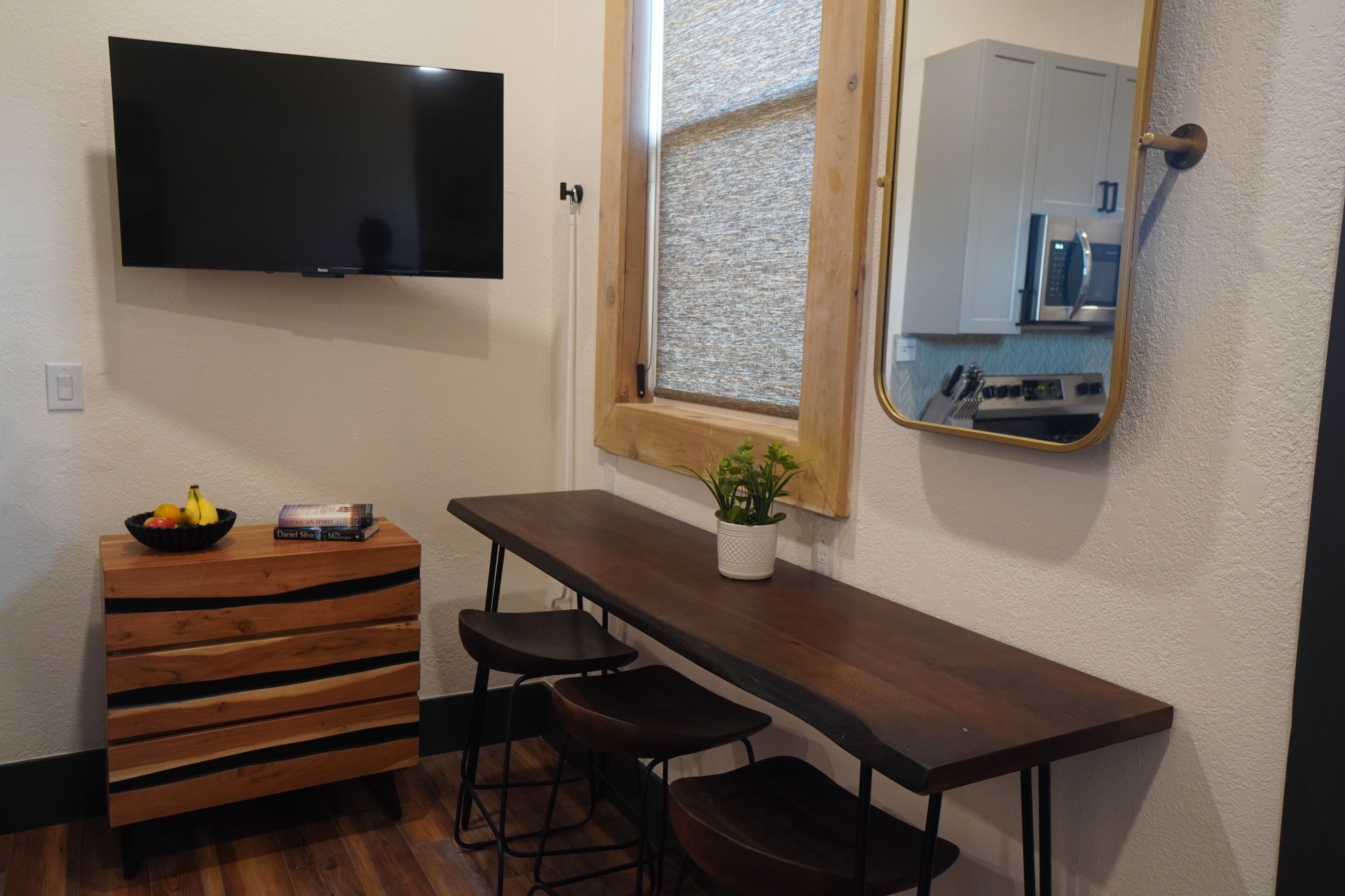 A modern kitchenette with a flat-screen TV, wooden bar stool seating, a small table with a plant, and a mirror reflecting the kitchen area.
