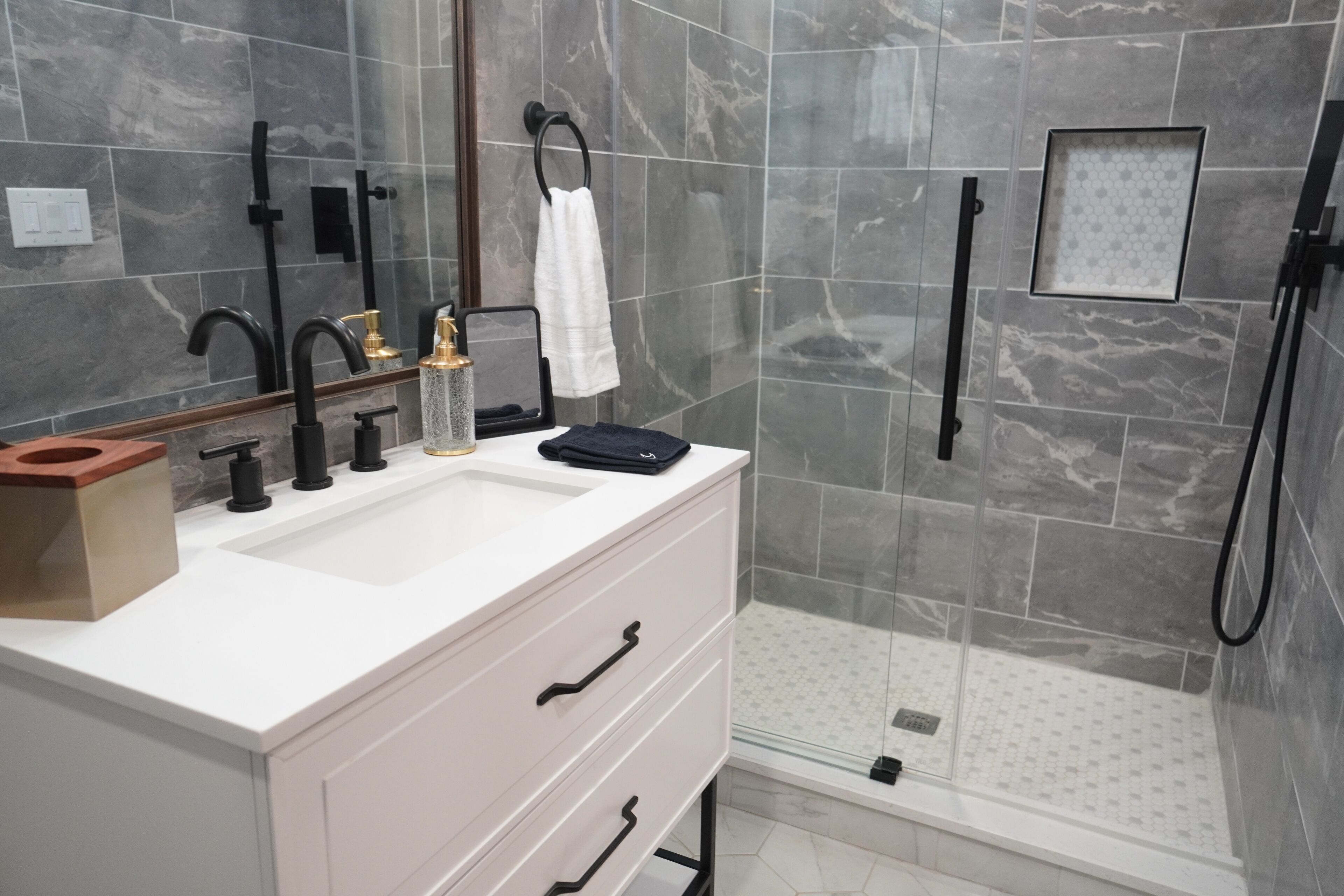 Un baño moderno con tocador blanco, grifos negros, cabina de ducha de cristal y zona de ducha con azulejos de mármol.