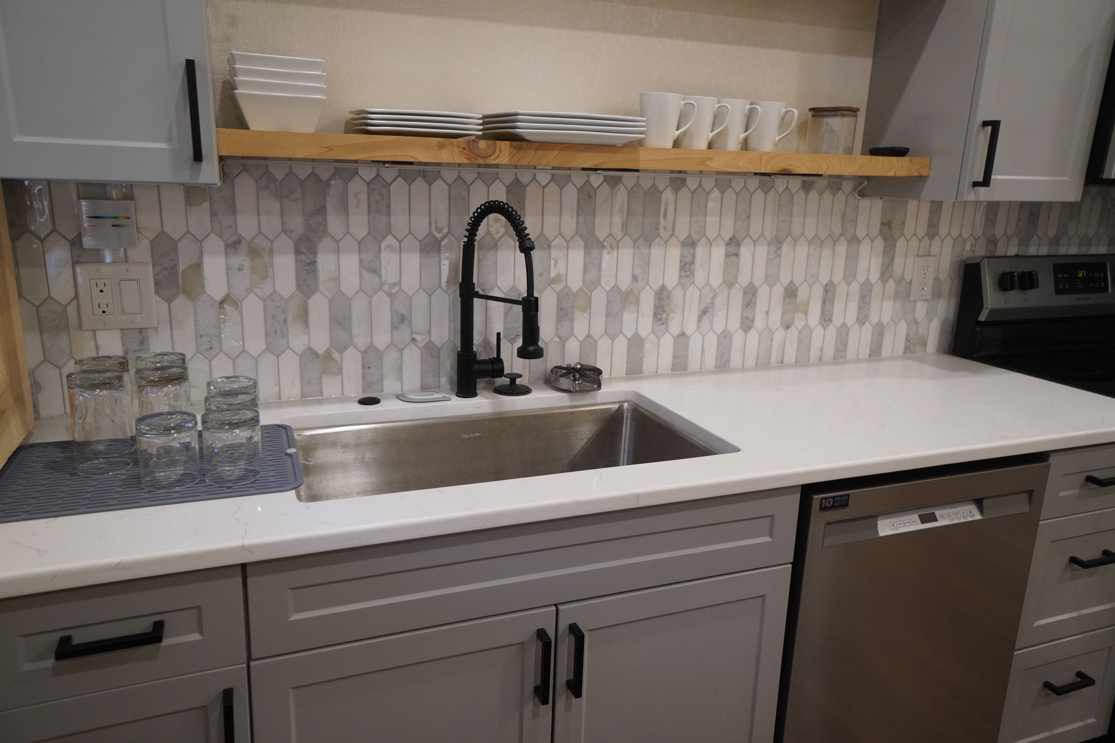 A kitchen with a white countertop, a stainless steel sink, and a dishwasher. There are cabinets with black handles and a wooden shelf above the sink.