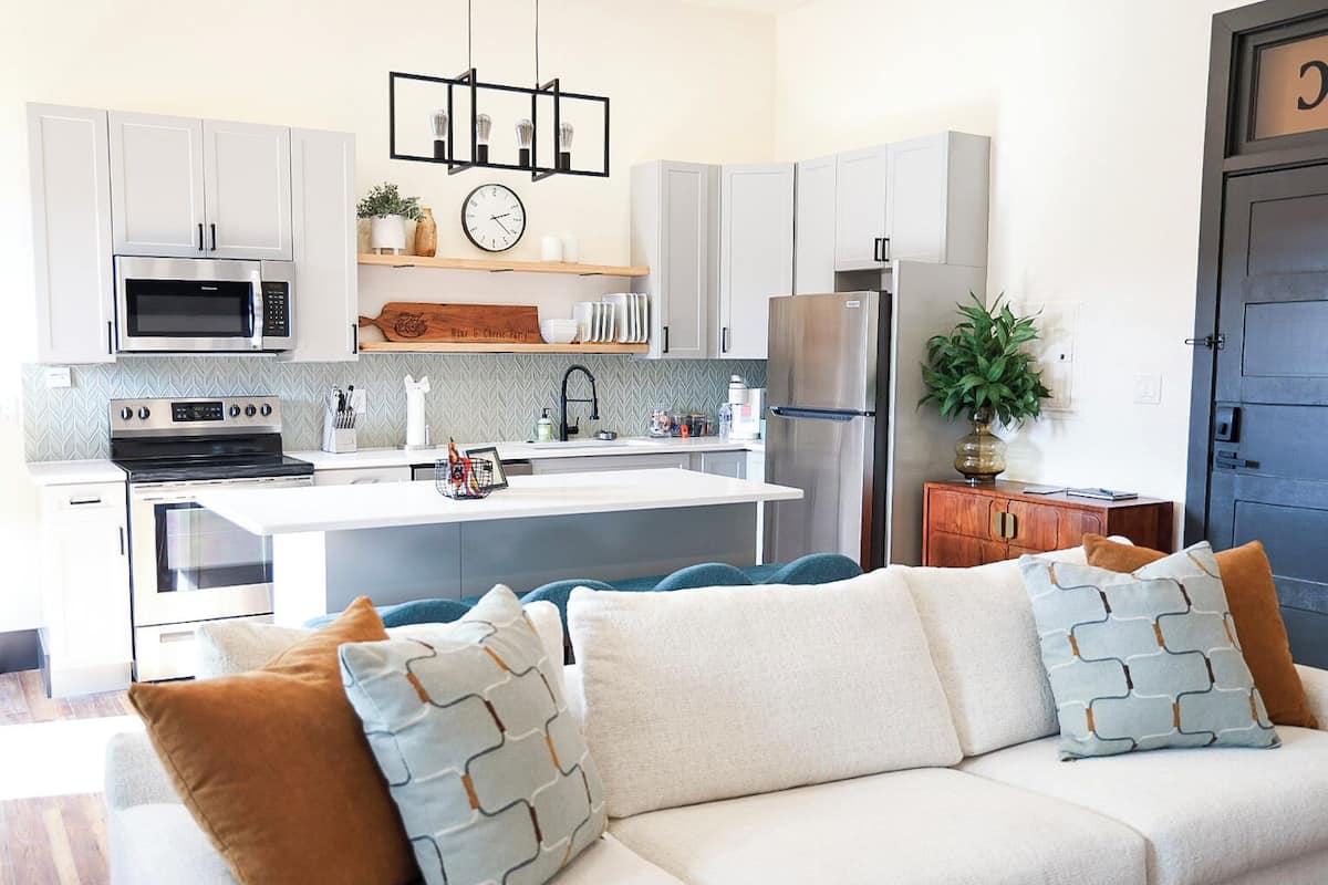 Una cocina moderna con horno empotrado, microondas y refrigerador, un comedor con una mesa blanca, y una sala de estar con sofá y cojines.