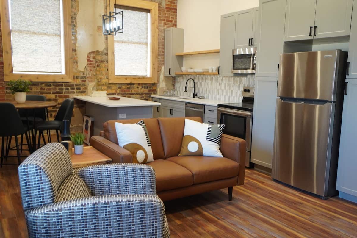 Una cocina moderna con un refrigerador de acero inoxidable, un rincón de comedor con un sofá marrón y sillas de mimbre, y una pared de ladrillo con ventanas.