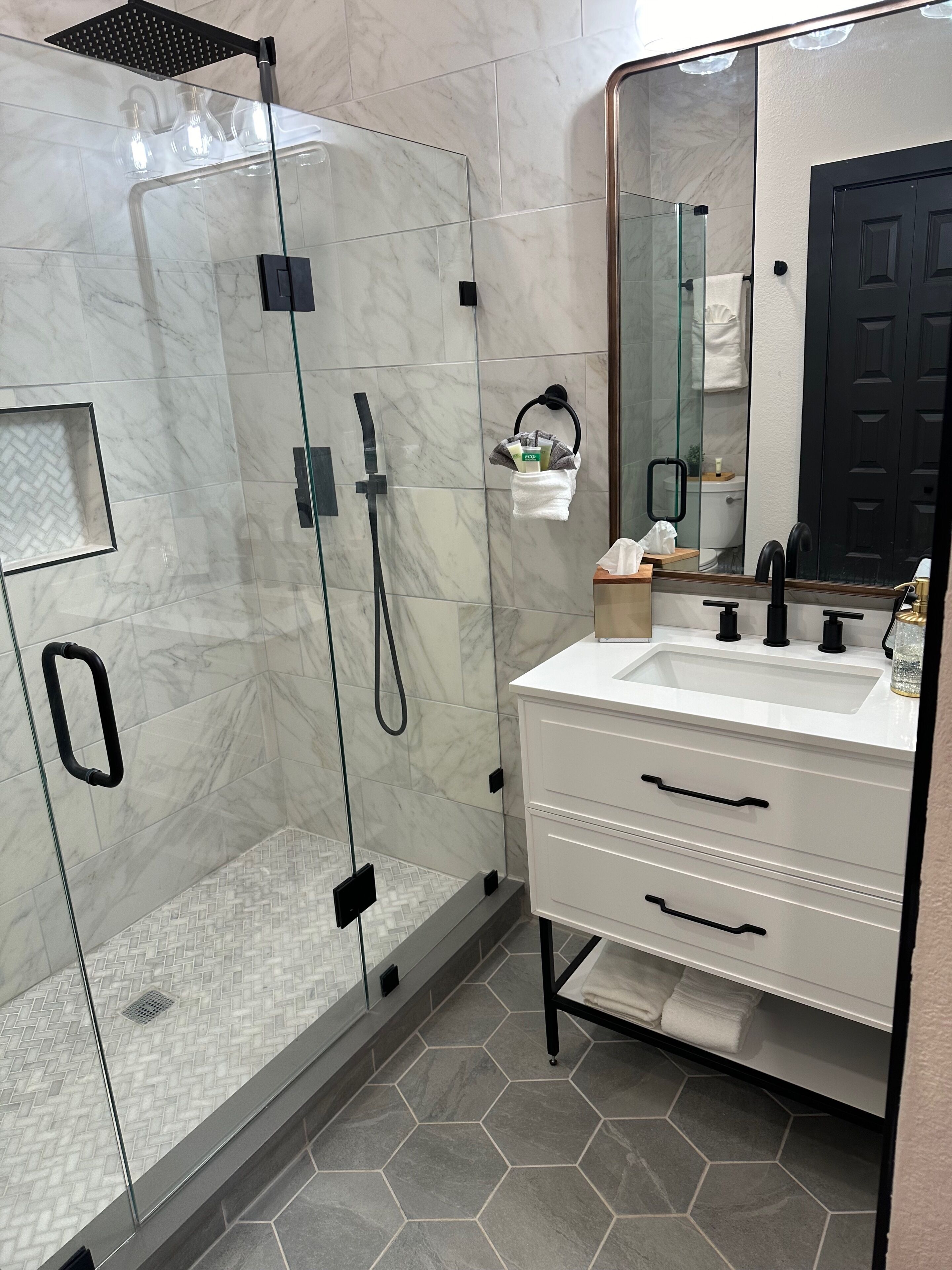 Een moderne badkamer met een douchecabine van glas, een wit badkamermeubel met zwarte handgrepen en een grote spiegel.