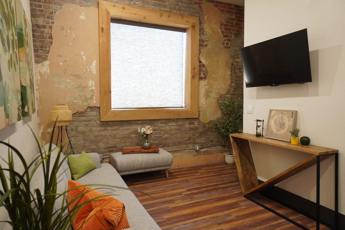 Una sala de estar con piso de madera, un sofá, una mesita, un televisor montado en la pared y una ventana con marco de madera.
