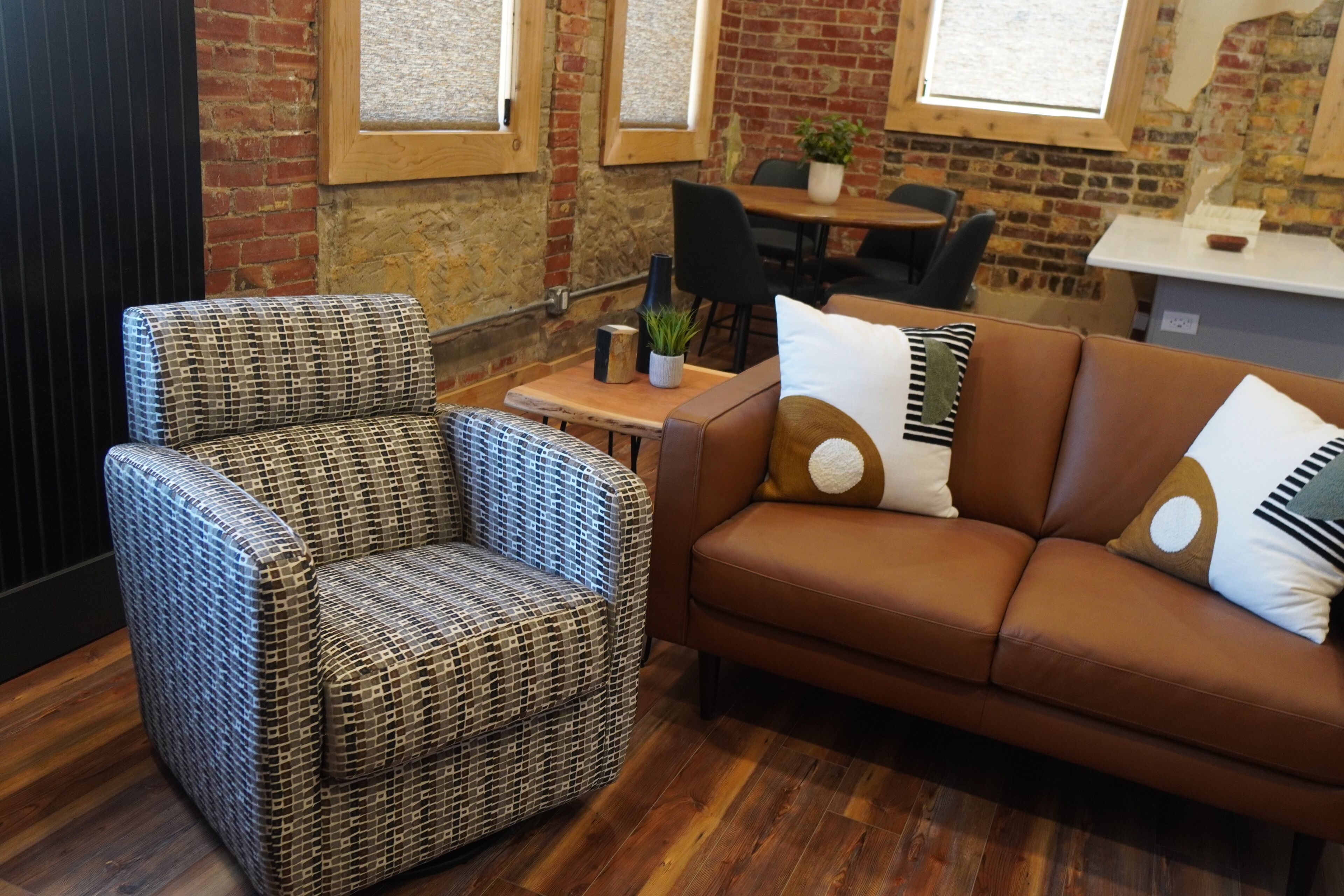 A room with a patterned armchair, a brown sofa with decorative pillows, a wooden table with a plant, and brick walls with windows.