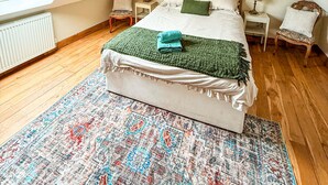A bedroom with a sloped ceiling, a large window, a bed with a green blanket, two bedside tables with lamps, and a patterned rug.