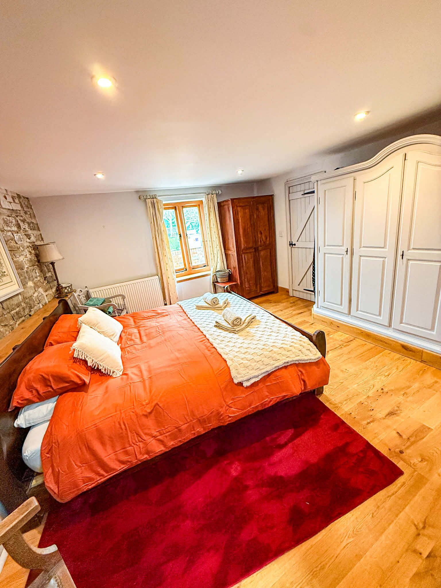 A bedroom with a large bed, a wooden floor, a stone wall, a window with curtains, and a wardrobe.
