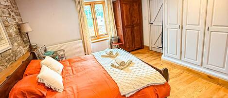 A bedroom with a large bed, a wooden floor, a stone wall, a window with curtains, and a wardrobe.