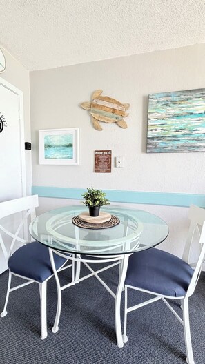 A round glass table with two white chairs, a potted plant, a framed picture, a wooden turtle decoration, and a 'House Rules' sign on a wall. - Coastal Oceanfront 1bd, pool and hot tub (Myrtle Beach)