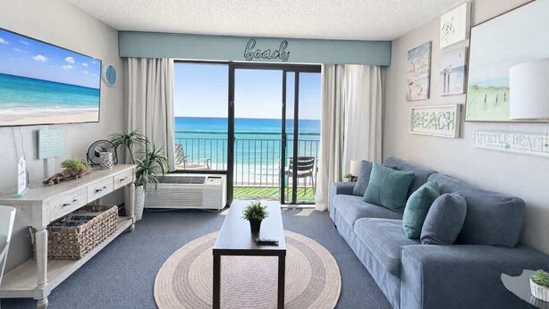 A living room with a view of the beach, a sofa, a coffee table, and a TV on the wall. - Coastal Oceanfront 1bd, pool and hot tub (Myrtle Beach)