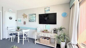 A room with a TV, a desk, a chair, a clock, and a plant. - Coastal Oceanfront 1bd, pool and hot tub (Myrtle Beach)