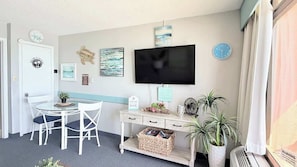 A room with a TV, a desk, a chair, a clock, and a plant. - Coastal Oceanfront 1bd, pool and hot tub (Myrtle Beach)