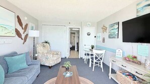 A living room with a sofa, armchair, coffee table, and a dining area with a table and chairs. - Coastal Oceanfront 1bd, pool and hot tub (Myrtle Beach)