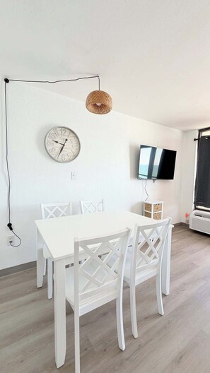 A minimalist dining area with a white table and chairs, a wall clock, a flat-screen TV, and a woven pendant light. - Awesome Oceanfront 1bd pool and hot tub (Myrtle Beach)
