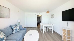 Ein Wohnzimmer mit einer Couch, einem Couchtisch, einem Esstisch mit Stühlen, einem an der Wand befestigten Fernseher und einer Tür zu einem weiteren Raum.