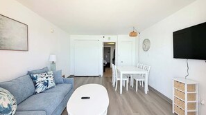 Ett vardagsrum med en soffa, ett soffbord, ett matbord med stolar, en tv som är monterad på väggen och en dörr som leder till ett annat rum.