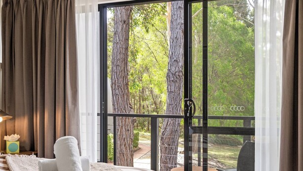 Une chambre à coucher comprenant un lit, une table de chevet, une lampe et une vue sur une terrasse et des arbres, visible à travers une porte-fenêtre coulissante.