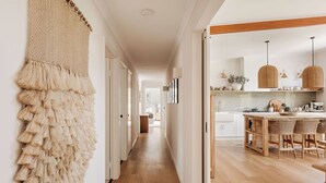 A hallway with a textured wall hanging, a wooden floor, and a kitchen area with hanging lights and a wooden table.