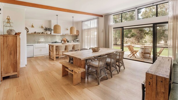 A spacious kitchen with a large dining table, wicker chairs, and a view of the outdoors through large windows.