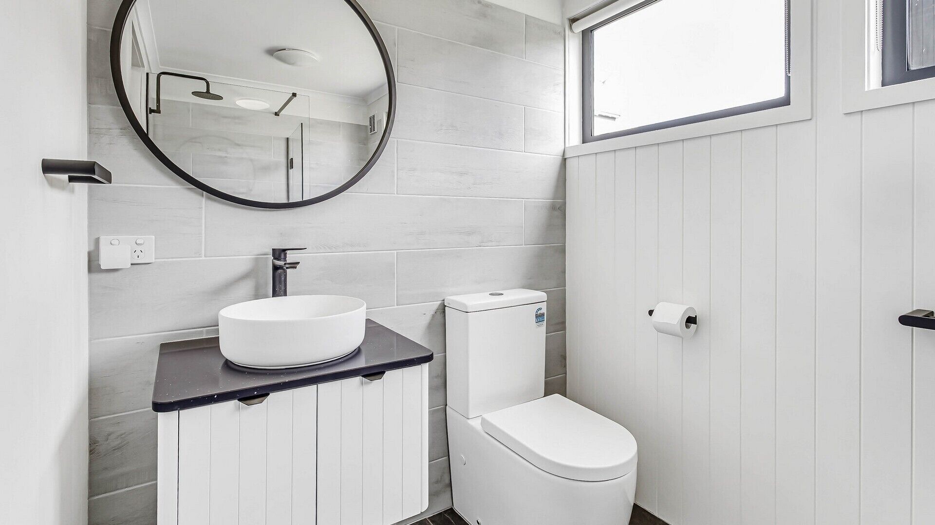 A modern bathroom with a round mirror, a white sink, a toilet, and a window.
