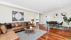 A modern living room with a brown leather sofa, a coffee table, and a dining area with a white table and black chairs. - Sasha's Cottage on the Hill | BBQ & Entertainment (Perth)