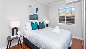 A bedroom with a bed, two bedside tables with lamps, a window with blinds, and a painting on the wall. - Sasha's Cottage on the Hill | BBQ & Entertainment (Perth)