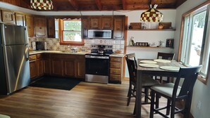 Eine Küche mit Holzausstattung, einem Edelstahl-Kühlschrank, einer Mikrowelle und einem Backofen. Ein Esstisch mit Stühlen und ein Fenster mit Blick auf Bäume.