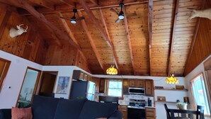 Ein rustikales Blockhaus-Interieur mit Holzbalken, einem Deckenventilator, einem aufgehängten Hirschkopf, einer Küche mit Geräten und einem Essbereich.