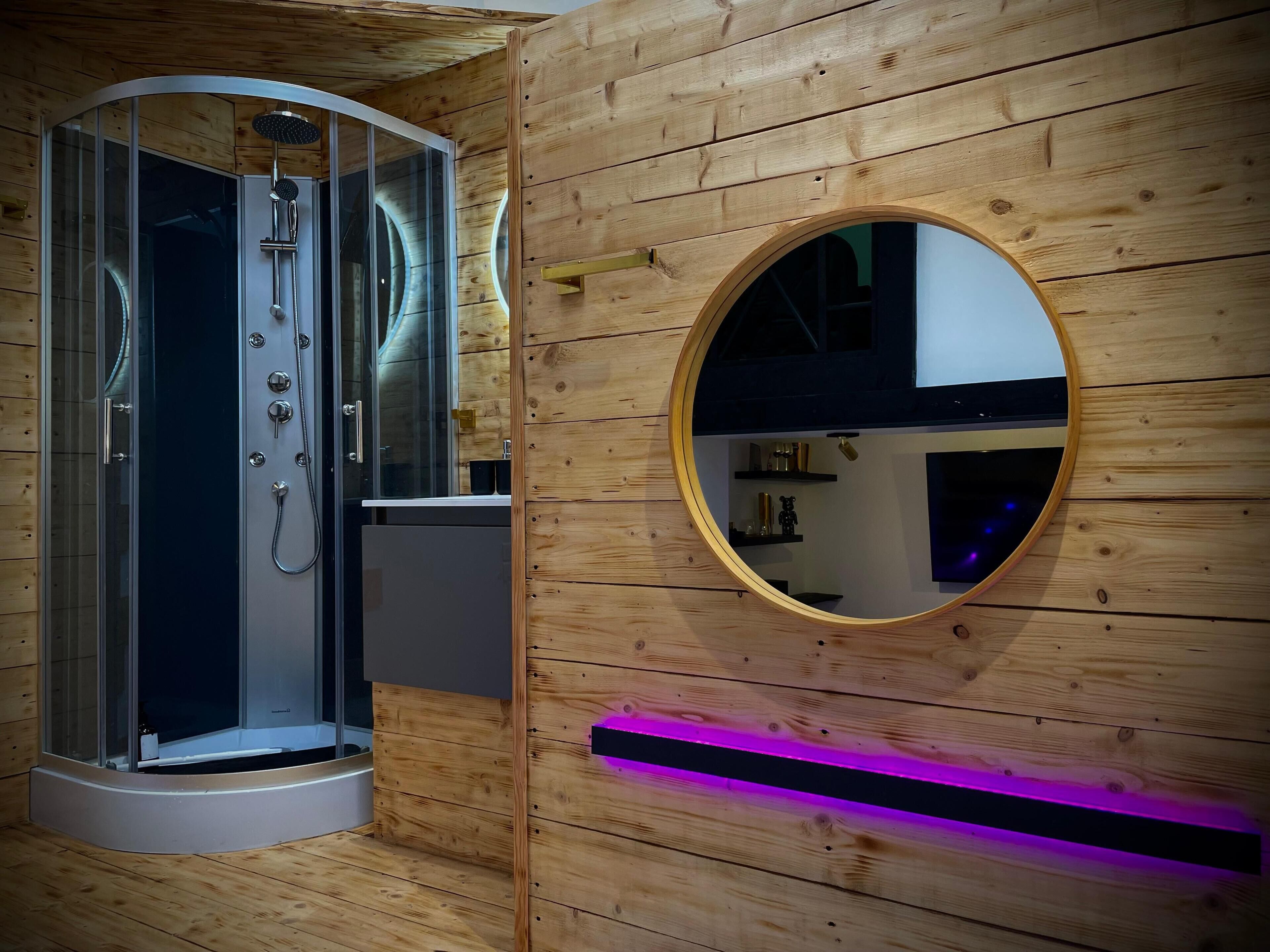 Une cabine de douche moderne dotée d’une porte en verre, d’un mur en bois orné d’un miroir rond et d’une étagère intégrée où sont disposés des objets décoratifs.