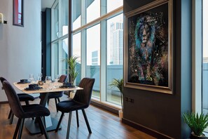 Uma sala de jantar moderna com mesa posta para seis pessoas, janelas amplas e um quadro abstrato emoldurado na parede.