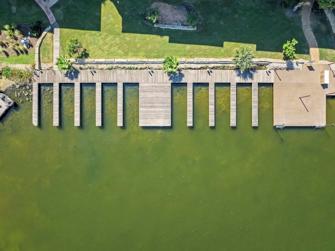 An aerial view of a dock extending into a body of water, surrounded by greenery and a paved walkway.