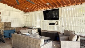 Un espace de vie extérieur abrité, doté de meubles en osier, d’un sol en pierre, d’un ventilateur de plafond et d’un téléviseur fixé au mur.
