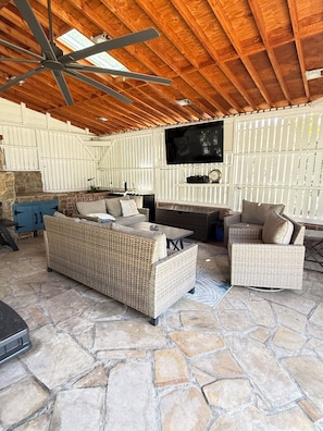 A covered outdoor living area with wicker furniture, a stone floor, a ceiling fan, and a mounted television. - Historic Pacific Beach Home with Putting Green, Jacuzzi, and Pool Table (San Diego)