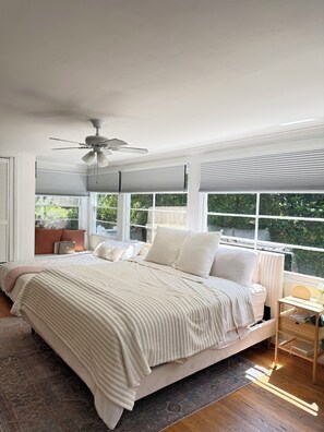 A bedroom with a bed, a ceiling fan, a window with blinds, a bedside table, and a view of greenery outside. - Historic Pacific Beach Home with Putting Green, Jacuzzi, and Pool Table (San Diego)