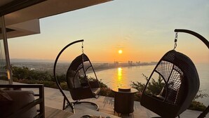 A modern outdoor seating area with two hanging chairs, a table, and a view of the sunset over a body of water. - Luxury villa with ocean view and private pool. Expectacular | 10 guests (Acapulco de Juárez)