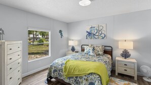 Une chambre avec une grande fenêtre, un lit recouvert d’une literie à motifs floraux, une commode, des tables de chevet avec des lampes et un ventilateur de plafond.