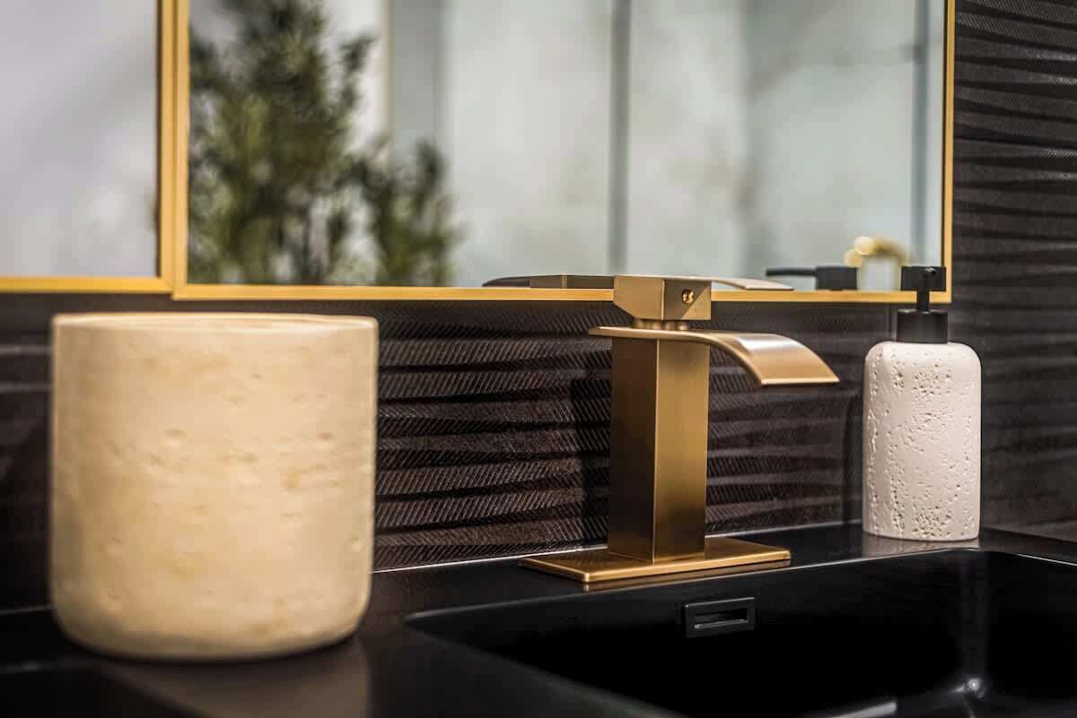 A bathroom with a black sink, a gold faucet, a beige vase, and a white textured container.