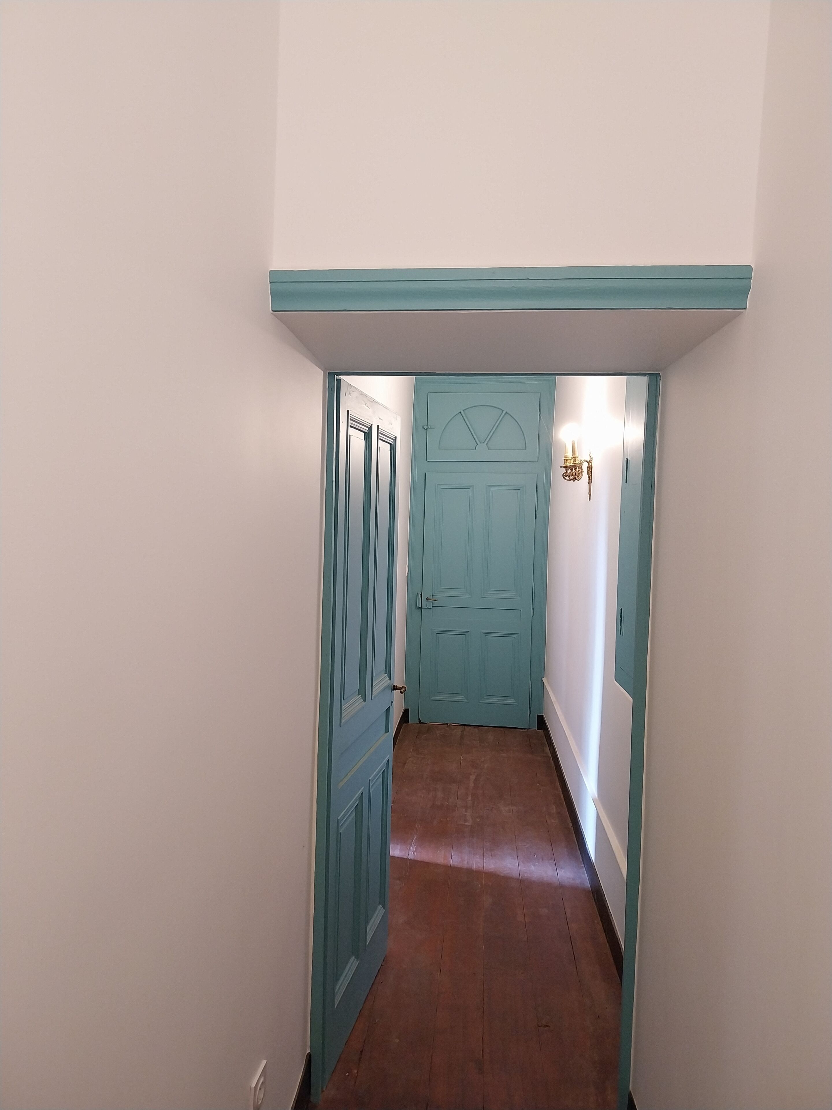 Um corredor estreito com piso de madeira que leva a uma porta verde-água com janela em formato de leque.