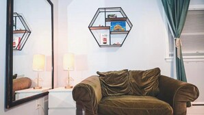 Una habitación con un sofá verde, un espejo, un estante hexagonal con libros, una lámpara y una ventana con cortinas.