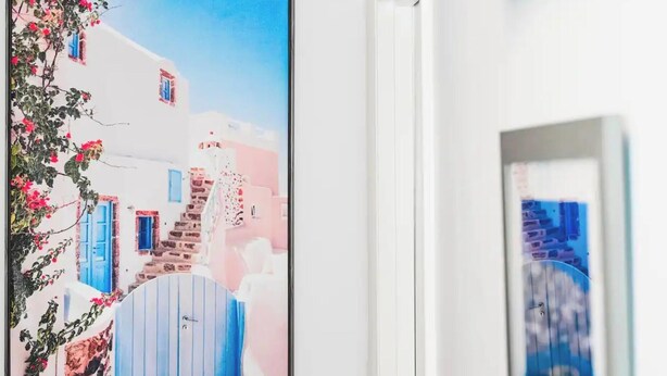 Un cuadro enmarco de un edificio blanco con puertas azules y paredes rosas, una escalera y flores rojas.