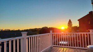Vista al atardecer desde un balcón con barandilla blanca, una terraza de madera y un horizonte urbano lejano.