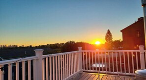 Ein Sonnenuntergangsblick von einem Balkon mit weißer Brüstung, einer Holzdielen-Terrasse und einer entfernten Stadtlandschaft.